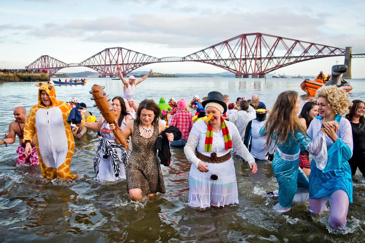 Loony Dook taking place at Queensferry, Edinburgh. Locals run into the sea on the first day of the year, some in fancy dress, with the Forth Rail Bridge behind.
